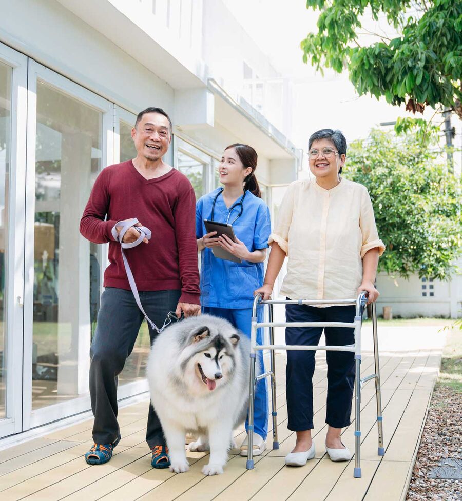 A caregiver with a tablet walks outside with an older couple, one using a walker, and their fluffy dog