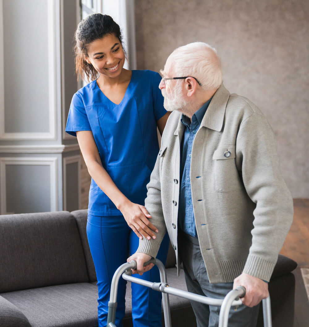 A caregiver in blue scrubs walks beside an older man using a walker at home